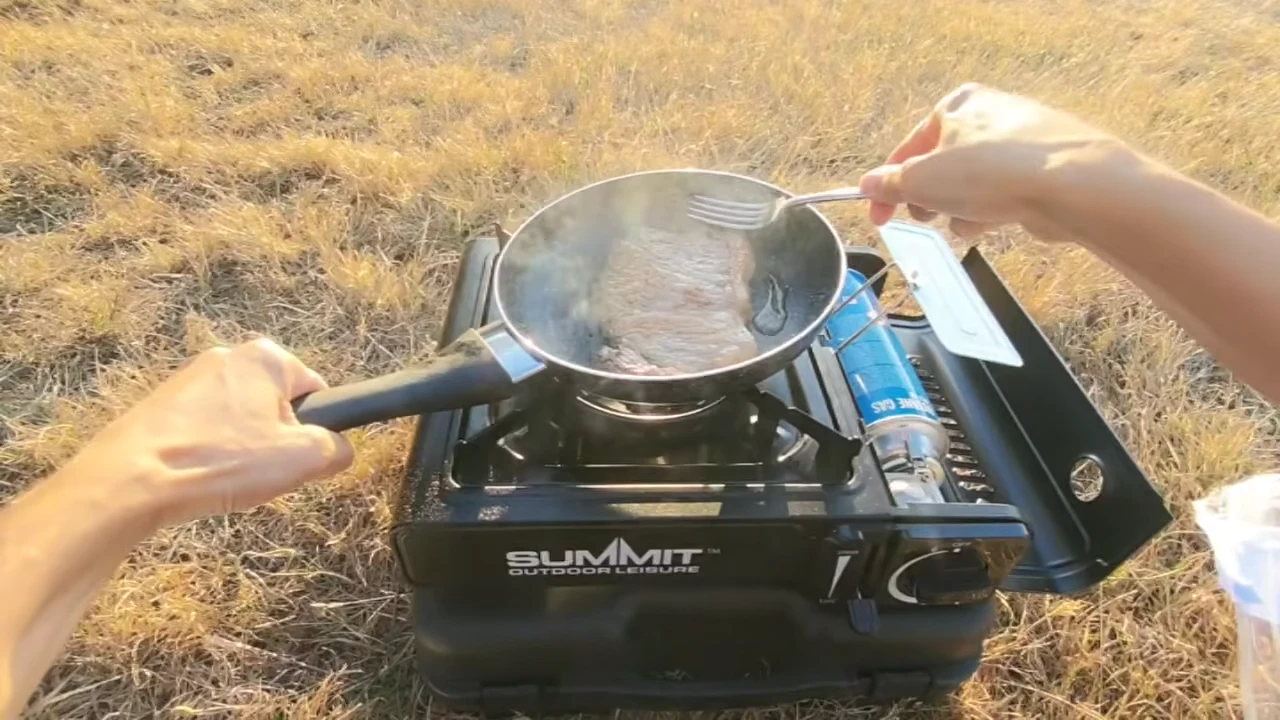 Hands stirring food in a pan on a portable single-burner stove on dry grass