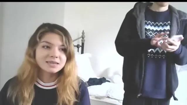 A woman seated in a bedroom-like room while Daniel stands partly in frame holding a phone