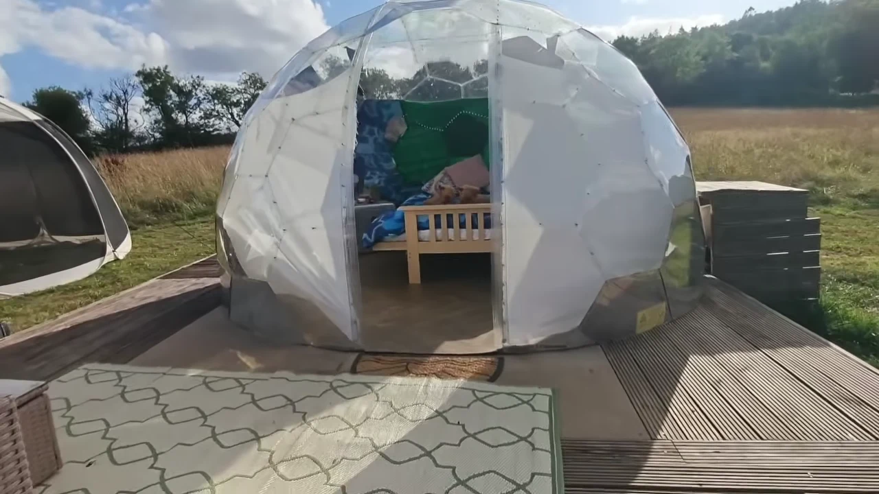 Transparent-panelled geodesic dome containing a bed, beside a dark shelter and outdoor furniture